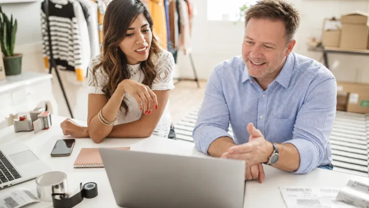 A boutique owner and a consultant review a laptop together at the shop counter, with shipping boxes and sales notes on the table.